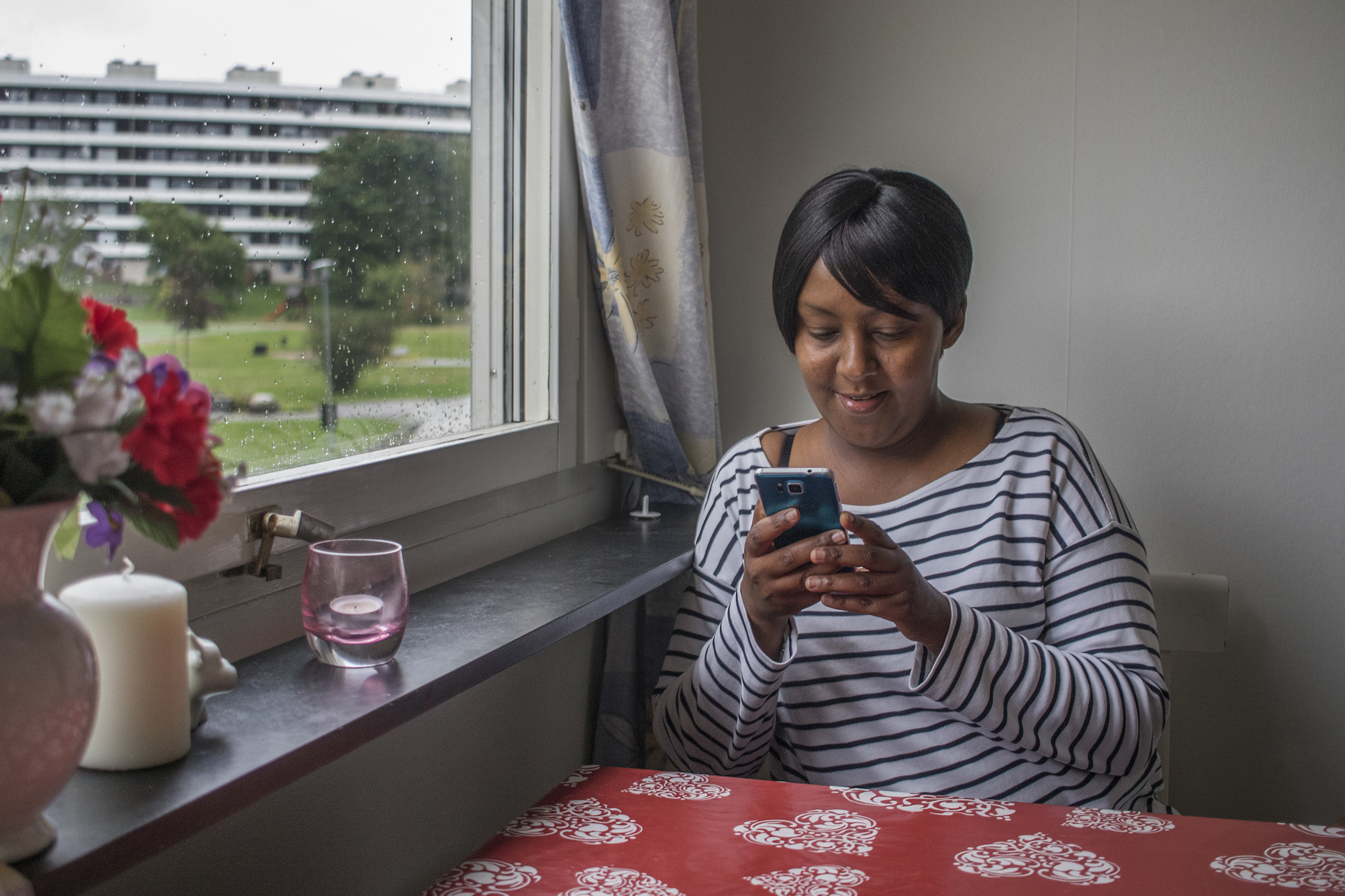 En person som sitter vid ett bord intill ett fönster och tittar på sin mobiltelefon.